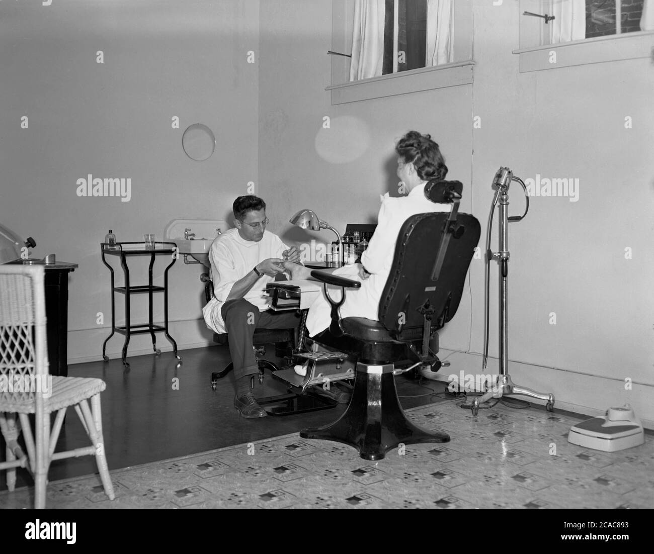 anni '40, storico, una signora seduta in una sedia girevole in un grande ambulatorio aperto o clinica che ha i suoi piedi trattati da un chiropodista maschile professionista che indossa una parte superiore medica bianca. Foto Stock