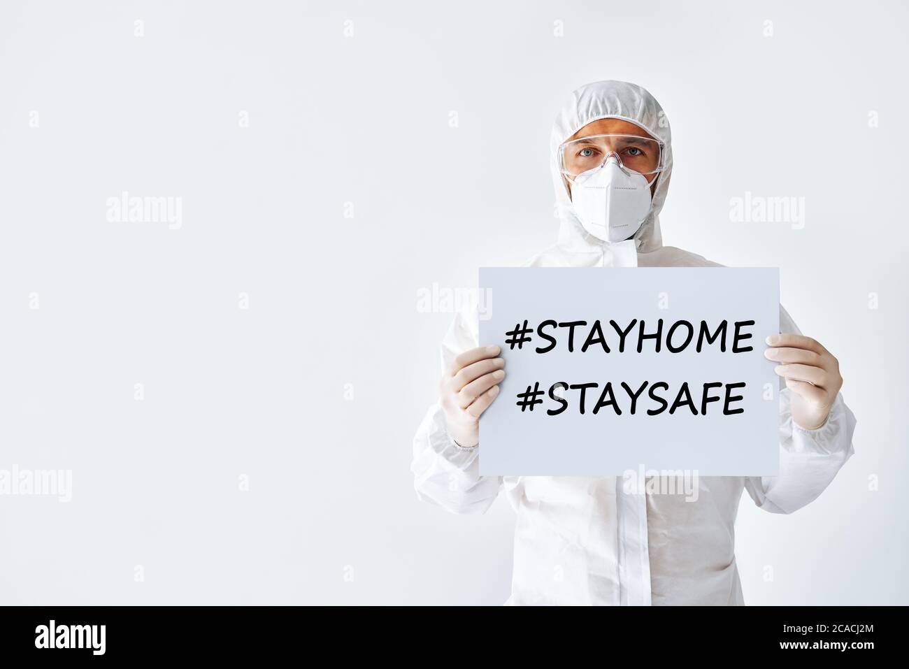 Dottore in tuta medica protettiva che mostra la targhetta vuota rimani a casa, rimani al sicuro su sfondo bianco Foto Stock