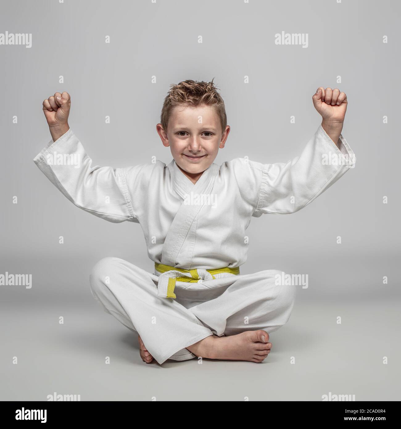 bambino allegro che pratica arti marziali seduto a terra con le braccia sollevate in un senso di vittoria. espressione felice. Foto Stock