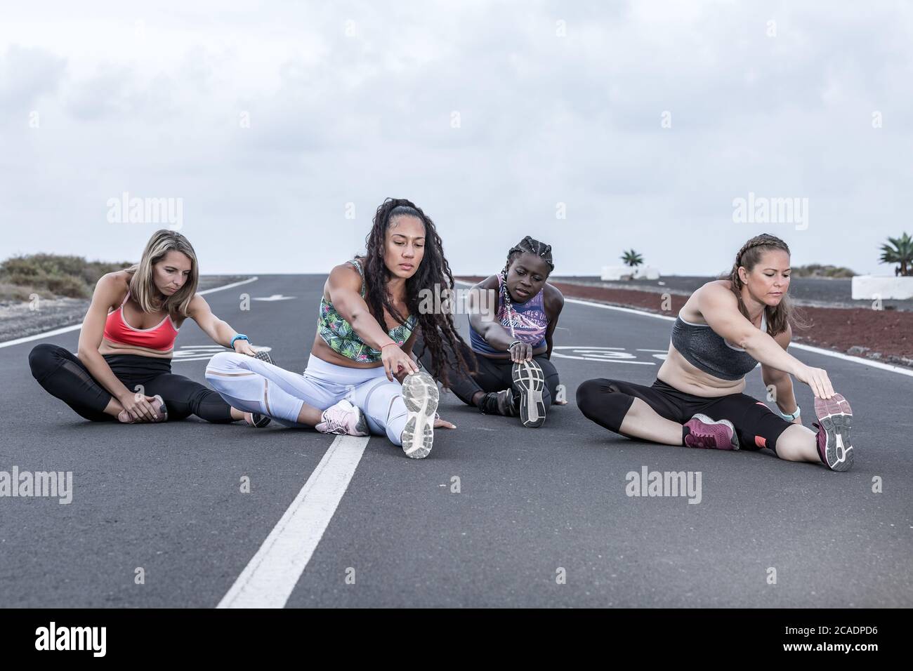 Atlete multietniche a corpo pieno in abbigliamento sportivo che si piega in avanti e tocca le dita mentre si siede su strada e si riscalda durante l'allenamento in gonna Foto Stock