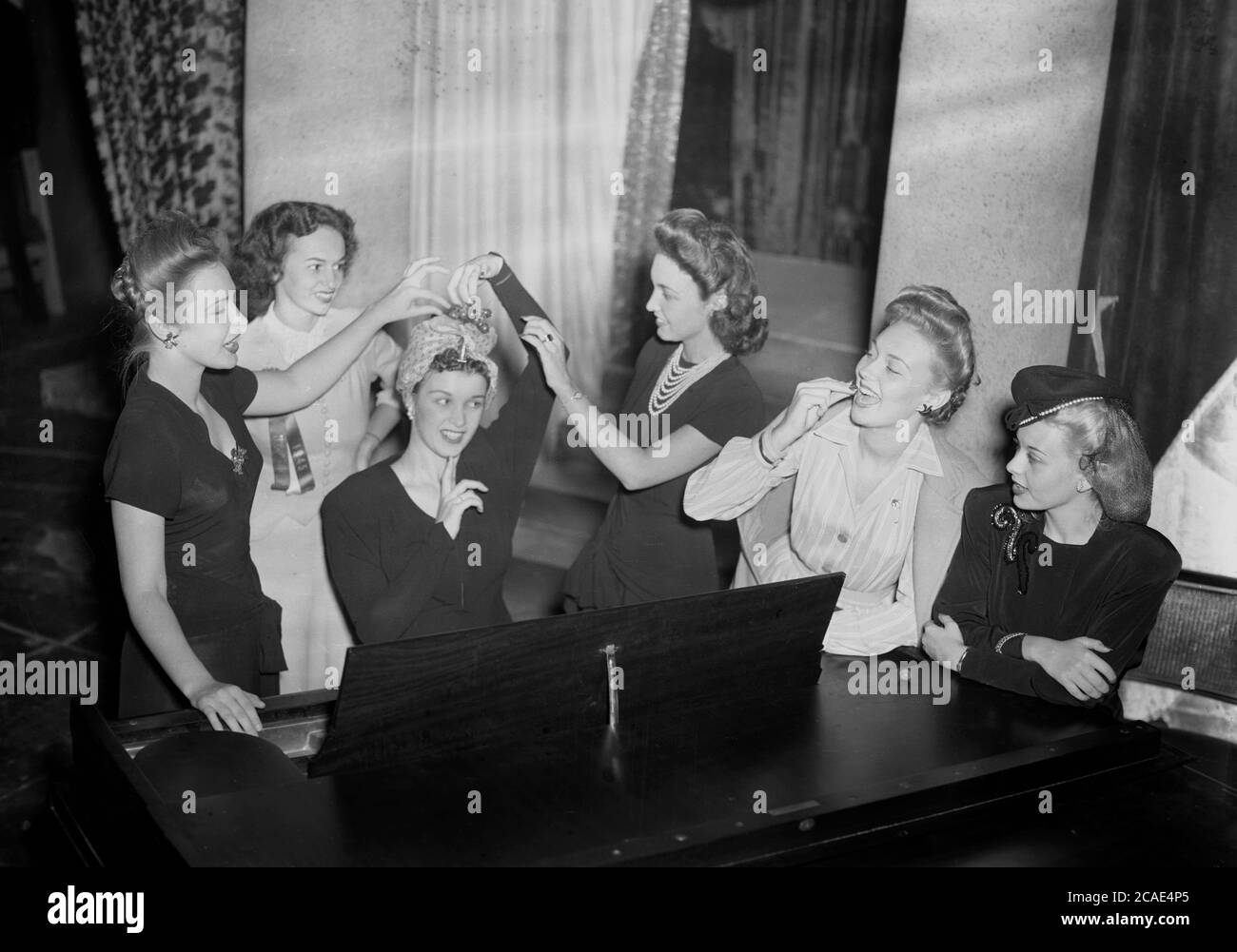 1945, storica, sei giovani donne attattive elegantemente vestite per una serata fuori in un salotto dell'hotel, divertendosi in piedi intorno al pianoforte dell'hotel che festeggia, con una delle giovani donne seduti al pianoforte che forse sta per suonare una canzone, USA. Foto Stock