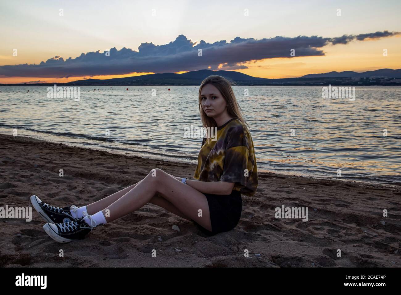 Una giovane ragazza è seduta su una spiaggia sabbiosa, con un paesaggio estivo drammatico, mare calmo e montagne dietro le quali il sole è scomparso Foto Stock