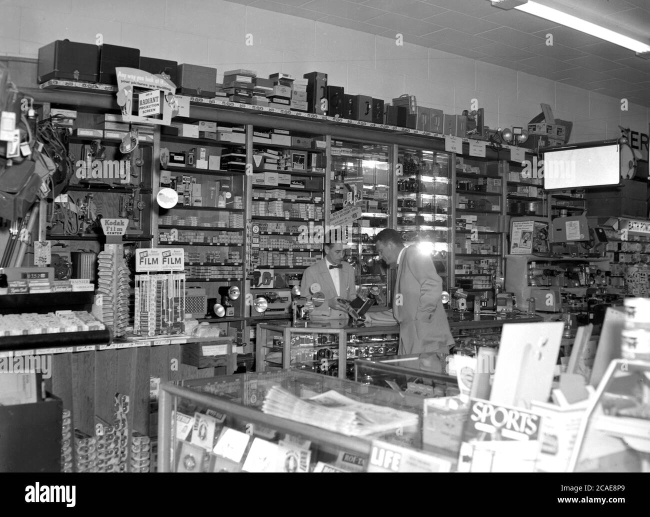 Anni '40, storico, al banco vendite, all'interno di un grande negozio fotografico, un rappresentante di vendita elegantemente vestito con un bow-tie, mostrando un cliente maschile una delle più recenti fotocamere, un modello con un 'soffietto' o lente estensibile, Stati Uniti. Le fotocamere con questo tipo di obiettivo pieghevole sono state normalmente utilizzate per la fotografia di medio formato dal fotografo più serio. Nella foto si possono vedere due nomi famosi di fotografia, Kodak e Polariod. I decenni successivi hanno visto l'ascesa di quello che si chiama "fotogiornalismo", la documentazione della vita ordinaria da parte della fotografia, Foto Stock