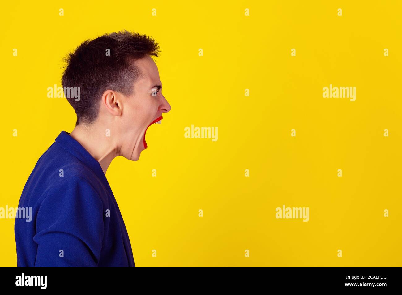 Vista laterale ritratto donna arrabbiata urlando, suono invisibile che esce dalla bocca aperta, isolato su sfondo giallo. Modello in camicia gialla e tuta blu Foto Stock