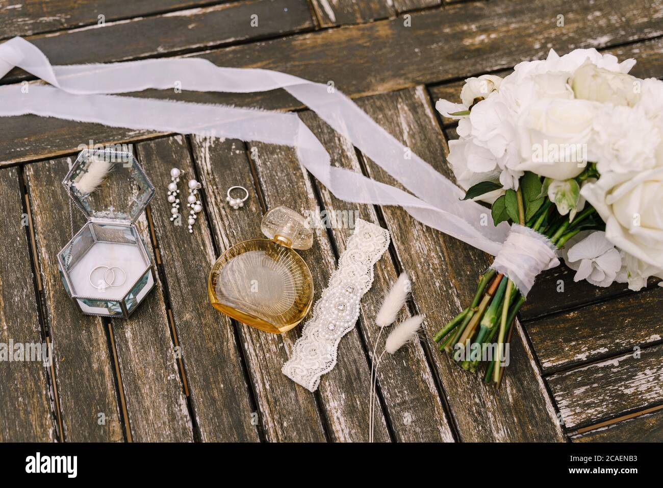 Un bouquet di rose bianche con un lungo nastro in sviluppo, un garter di apertura, una bottiglia di profumo, un anello con pietre, orecchini e anelli di nozze in un Foto Stock