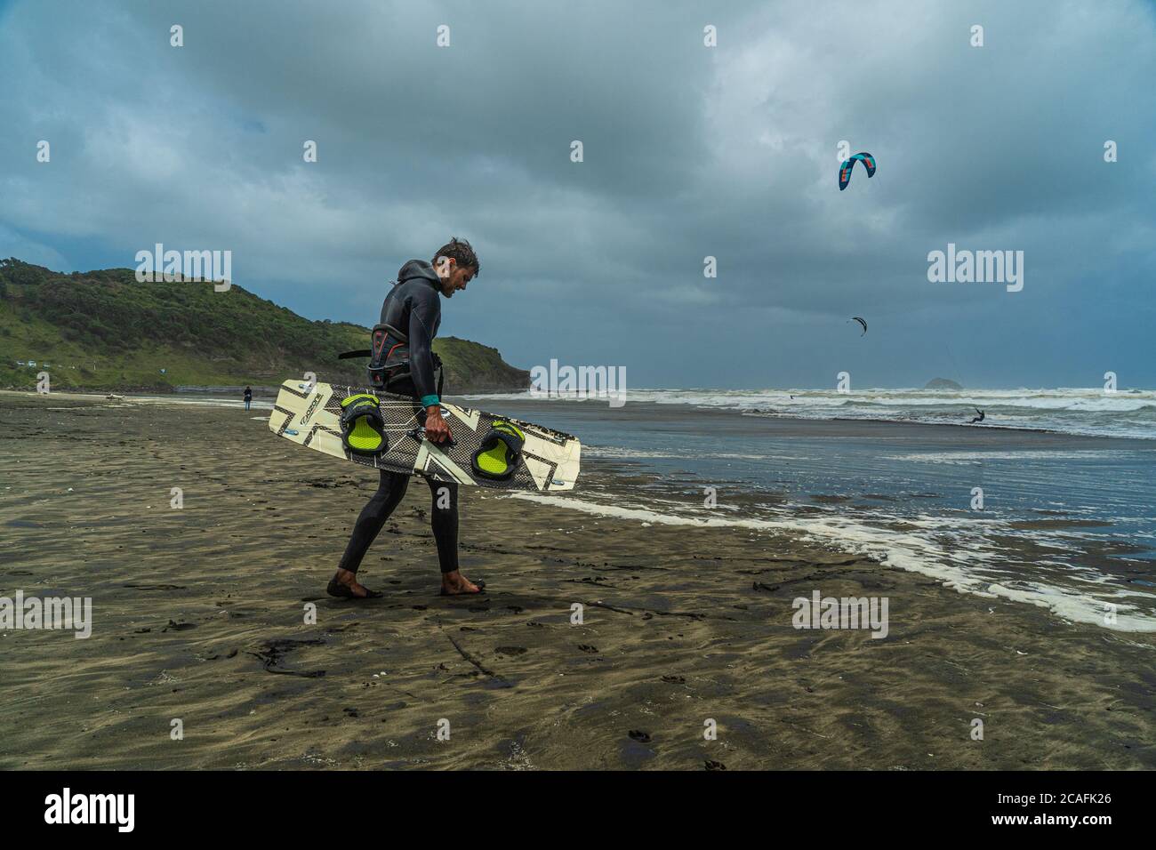 Un kitesurfer maschile in una muta porta il suo kiteboarding attraverso una spiaggia tempestosa. Un kitesurfer esegue un salto in background. Foto Stock