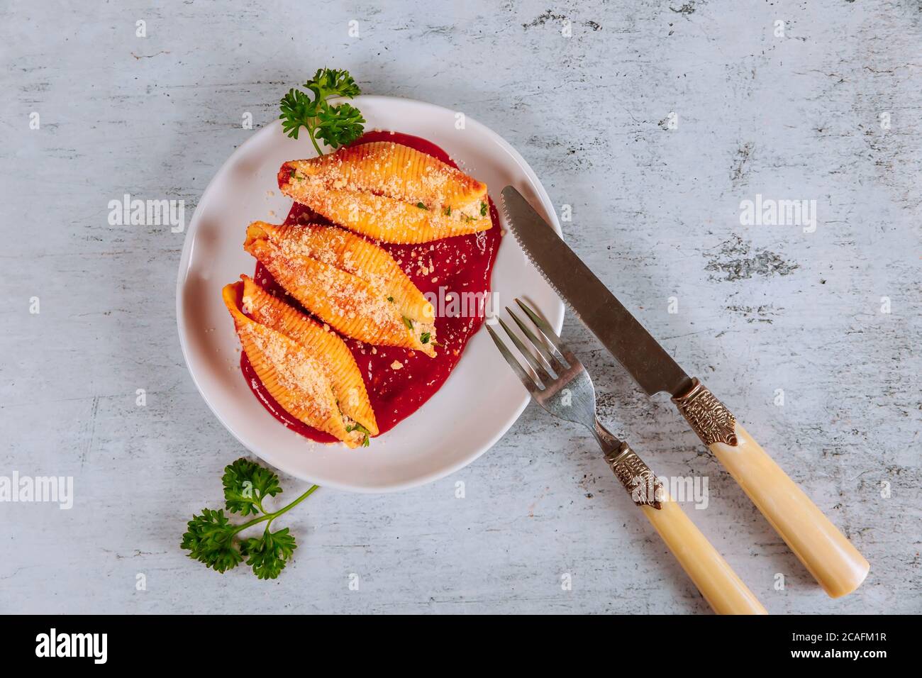 Conchiglie di pasta italiana farcite con ricotta, carne e parmigiano sul piatto con salsa di pomodoro, coltello e forchetta. Foto Stock