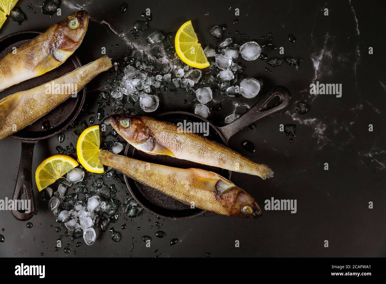 Pesce intero crudo con ghiaccio e limone su tavola nera. Vista dall'alto. Foto Stock