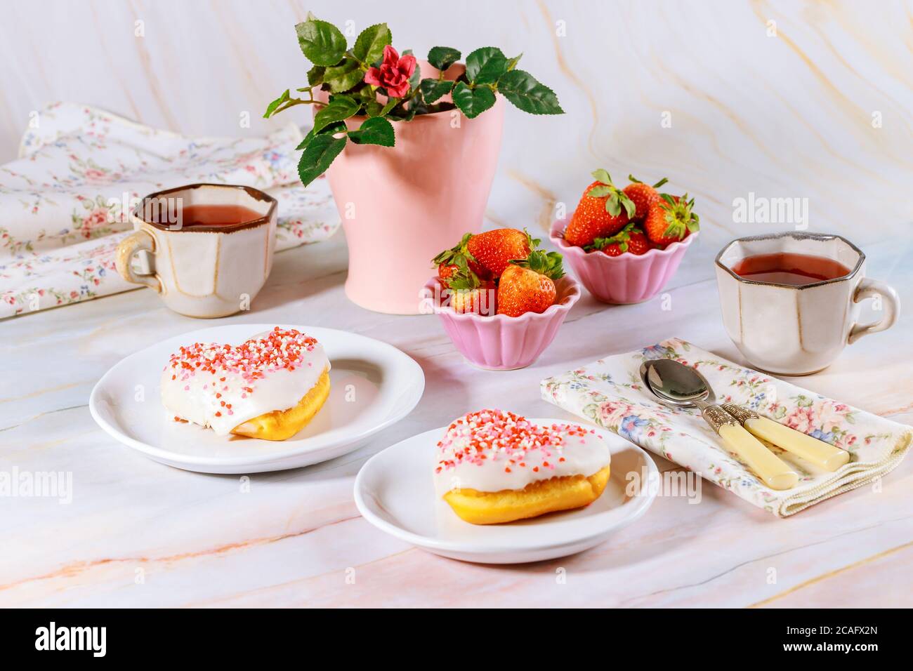 Due ciambelle dolci e rosa a forma di cuore con smalto bianco e cospargere sul tavolo da festa con tè, fragole e rosa. Concetto di San Valentino. Foto Stock
