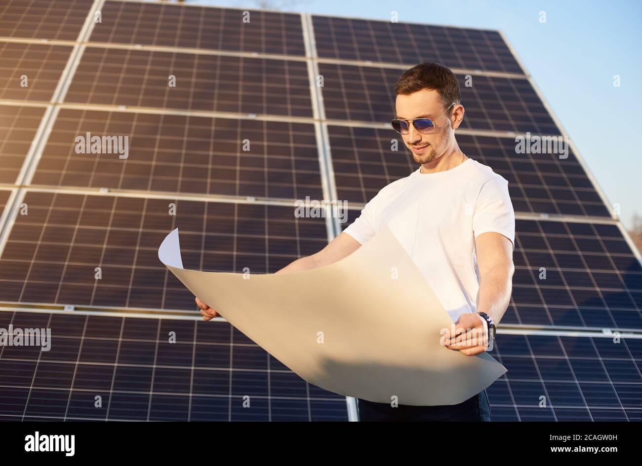 Giovane uomo d'affari sta guardando in un piano di carta bianca in piedi da un grande pannello solare, il sole splende brillantemente sul viso, concetto eco-friendly Foto Stock