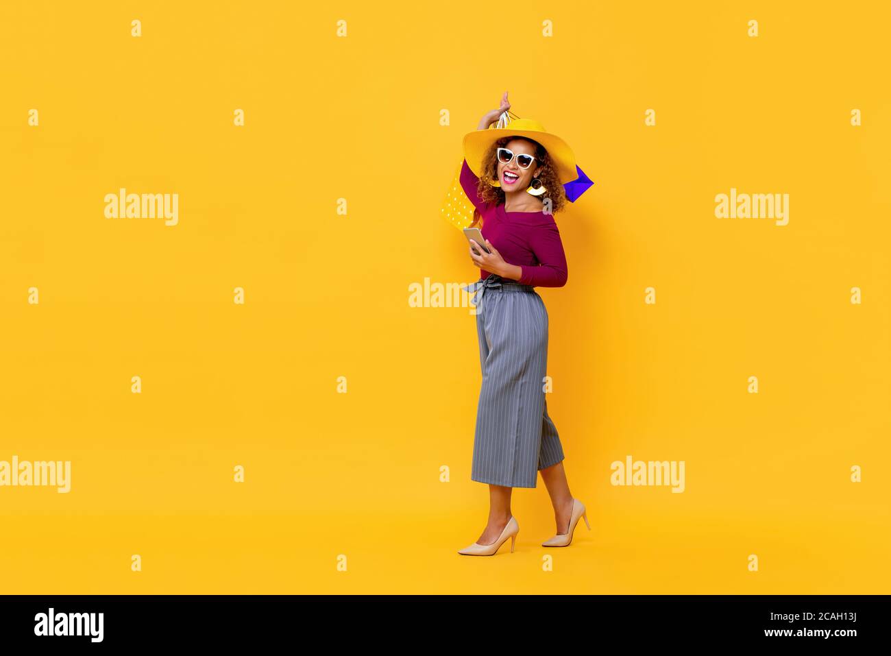 Ritratto a lunghezza intera di felice giovane attraente donna afroamericana sollevare borse per la spesa e tenere il telefono cellulare in studio isolato sfondo giallo Foto Stock