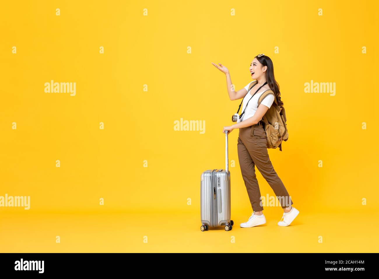 Ritratto a lunghezza intera di bella giovane donna turistica asiatica sorridente con la borsa del trolley che guarda e la mano di apertura per lo spazio di copia isolato su yello colorato Foto Stock