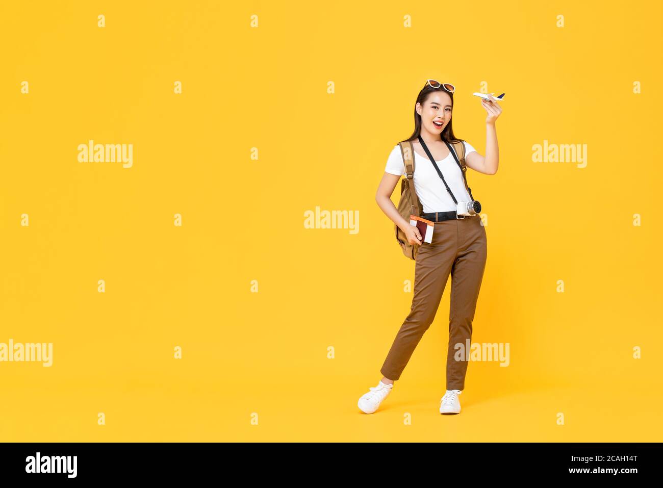 Sorridente giovane bella donna turistica asiatica tenendo modello aereo e. passaporto isolato su sfondo giallo studio Foto Stock