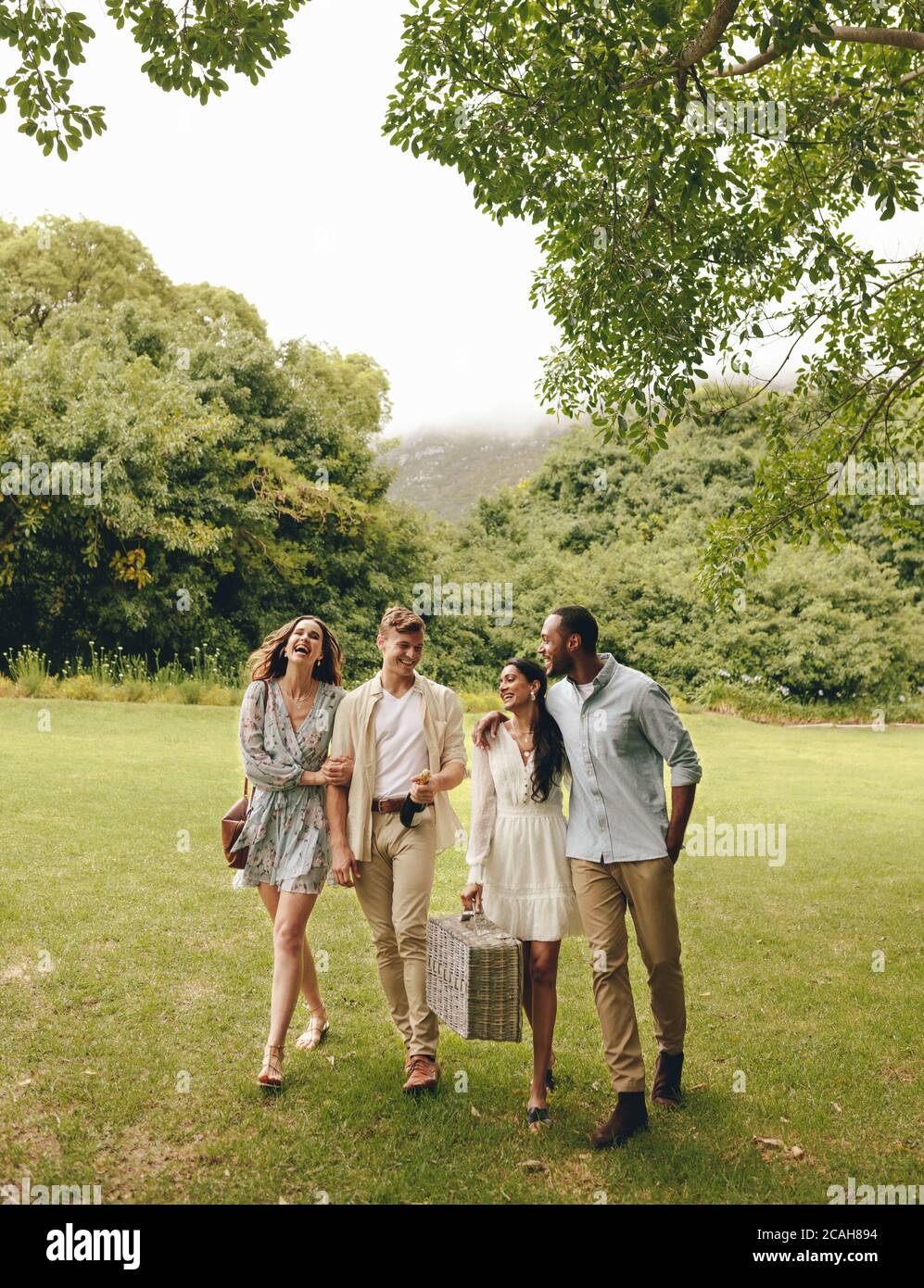 Due coppie che vanno al picnic nel parco cittadino. Amici che camminano insieme e sorridono in giardino. Foto Stock