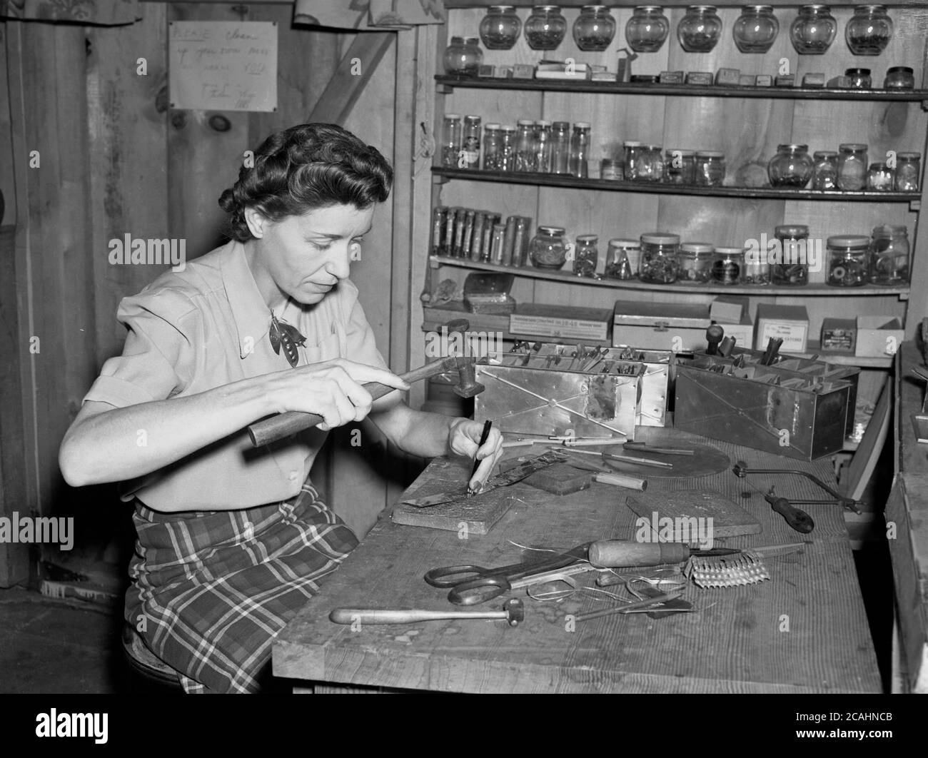 circa anni '40, storico, una signora in una piccola capanna di legno o capannone seduto a una panchina di quercia facendo intricate metallizzazioni a mano utilizzando un martello e un piccolo punzone utensile. Foto Stock