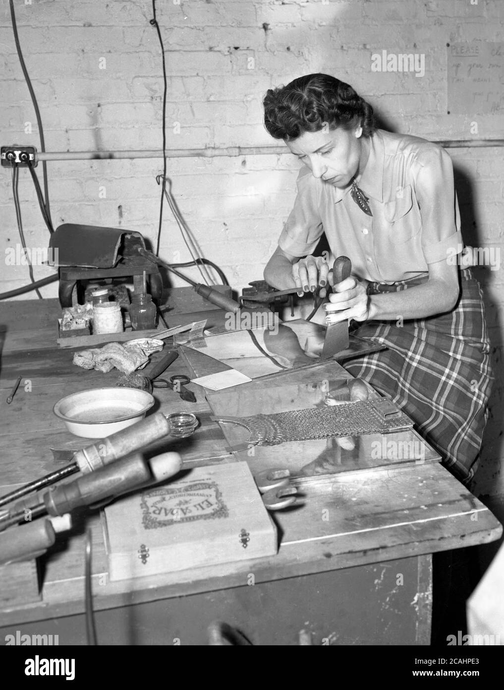 circa anni '40, storico, una signora in una piccola officina artigianale seduta su una panchina di quercia facendo intricate metallizzazioni a mano utilizzando un martello e un piccolo punzone. Foto Stock