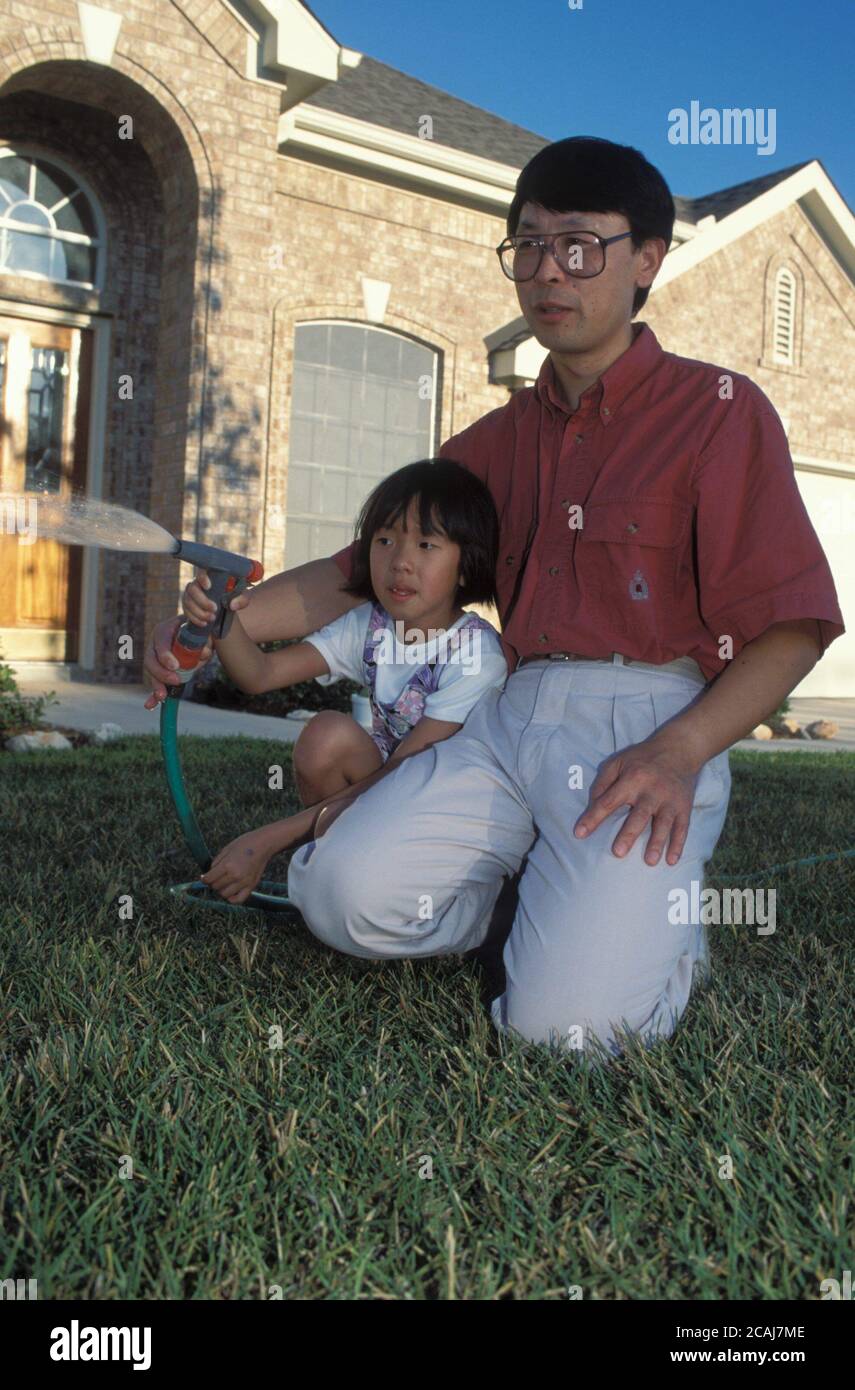 Padre e figlia cinesi-americani si mettono davanti a una nuova e attraente casa in mattoni mentre annaffiano il prato ad Austin, Texas, Stati Uniti. ©Bob Daemmrich Foto Stock