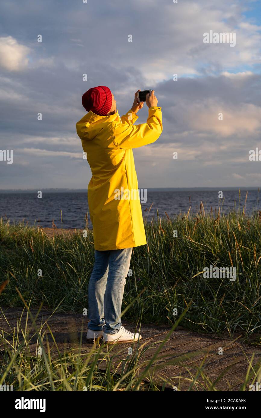 Uomo con impermeabile giallo indossa un cappello rosso in piedi sulla spiaggia su un sentiero in legno, guarda il cielo nuvoloso drammatico e scatta foto sullo smartphone. Stagione autunnale. Foto Stock
