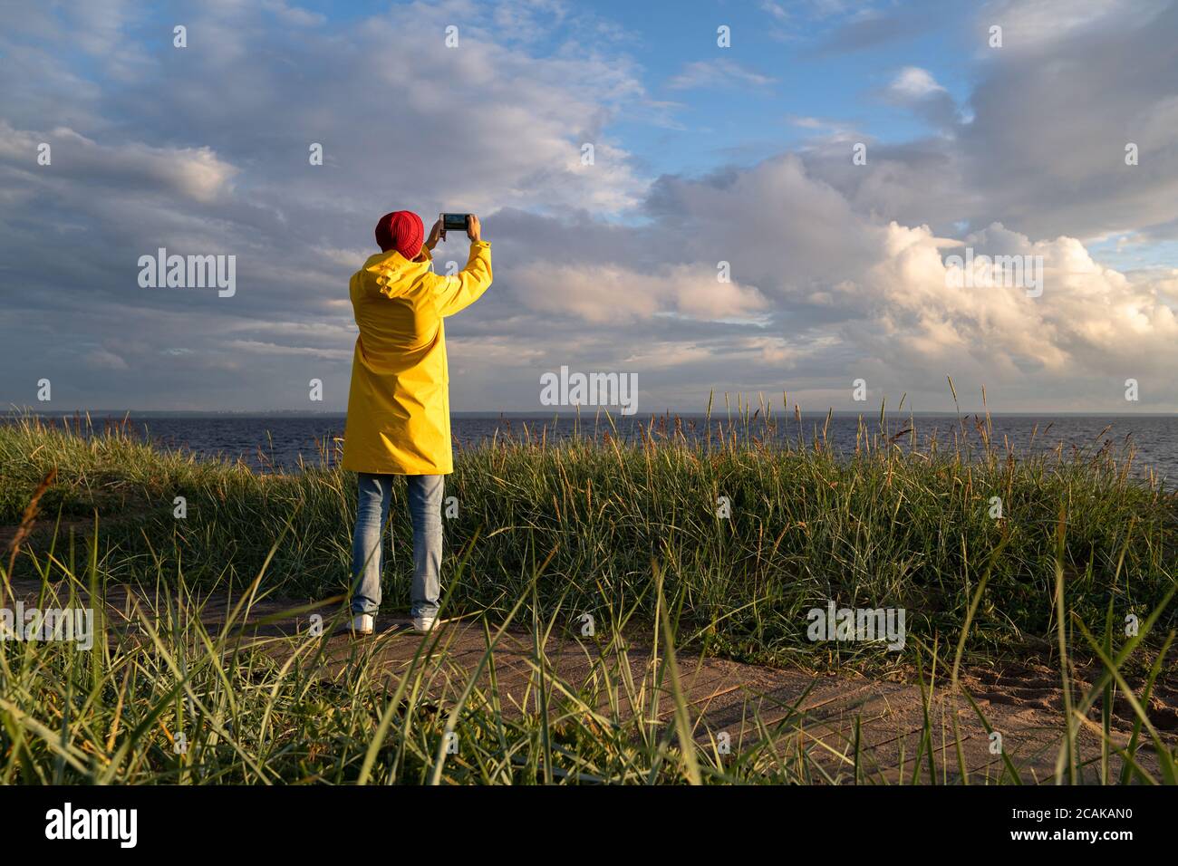 Uomo con impermeabile giallo indossa un cappello rosso in piedi sulla spiaggia su un sentiero in legno, guarda il cielo nuvoloso drammatico e scatta foto sullo smartphone. Stagione autunnale. Foto Stock