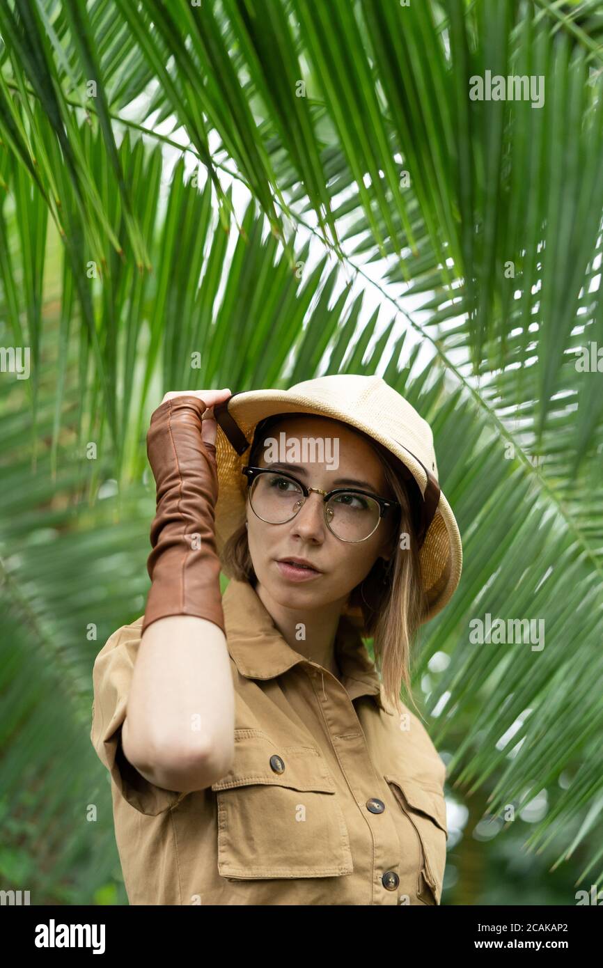 Donna botanica vestita in stile safari in serra. Naturalista in abiti cachi, guanti di lievito nella foresta pluviale circondati da palme. Giungla turistico Foto Stock