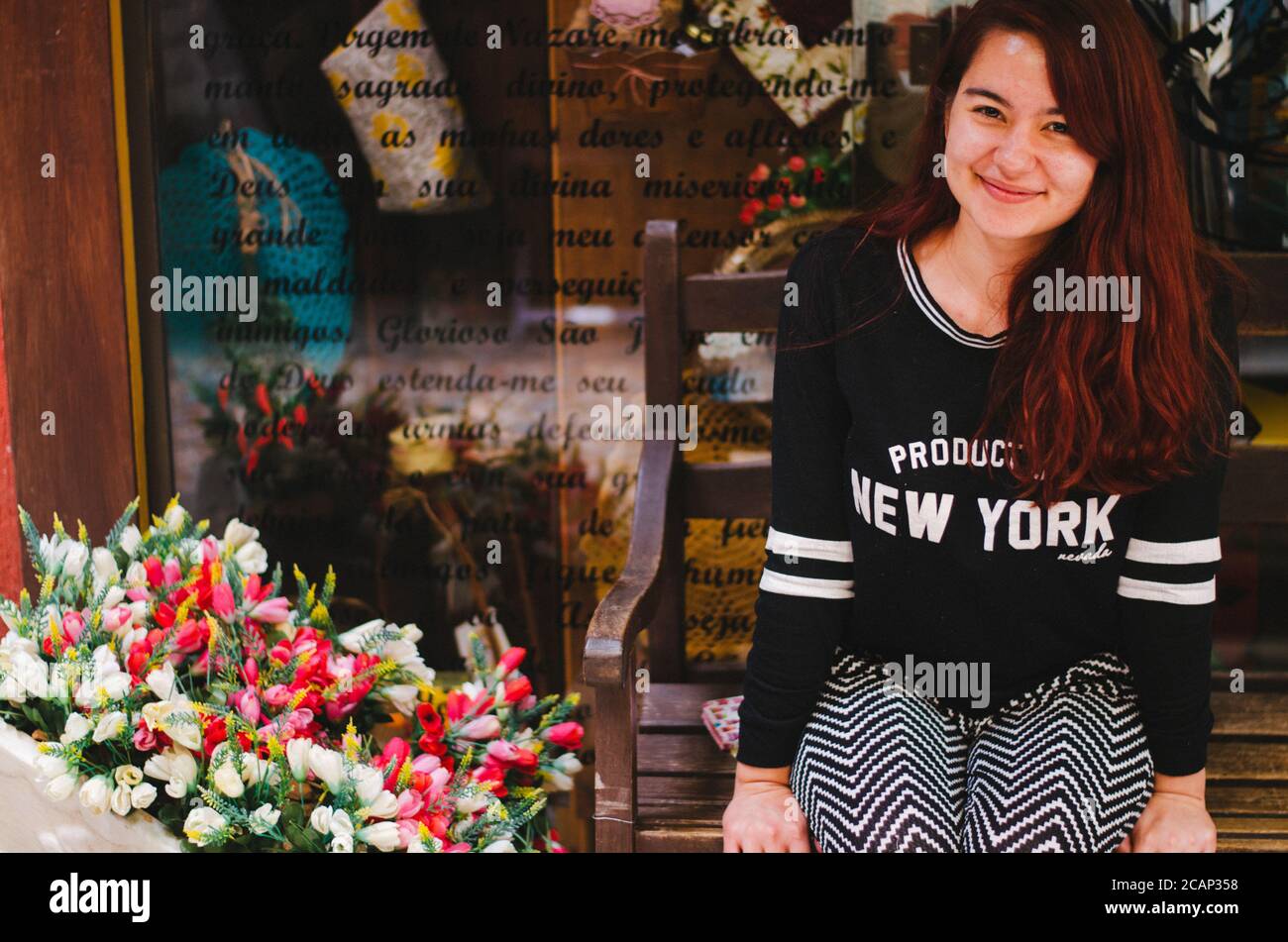 Un giovane adulto rosso testa brasiliano sorridente alla macchina fotografica mentre seduto in una panchina di legno accanto ad una disposizione dei fiori e dietro un negozio display con testo w Foto Stock