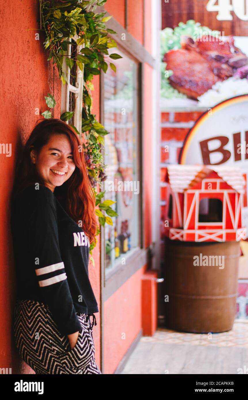 Un giovane adulto rosso testa brasiliano sorridente alla macchina fotografica mentre in posa accanto a un negozio con pareti rosse e. un cestino a forma di graziosi coloni rossi Foto Stock