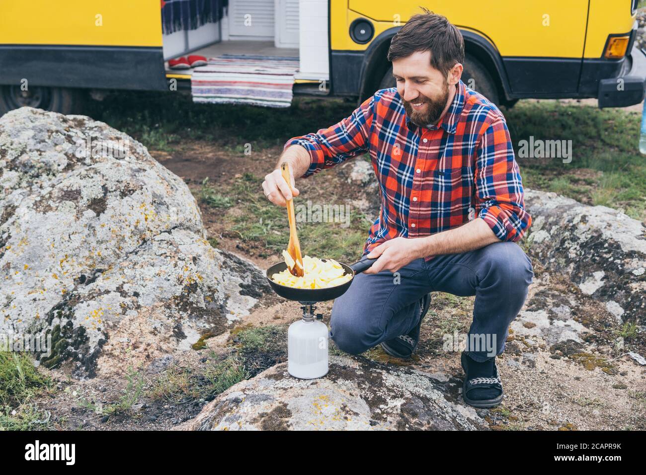 Giovane uomo che cucina in campervan mentre si soggiorna in campagna. Campeggio off-grid auto-costruito in campeggio nella natura selvaggia. Foto Stock