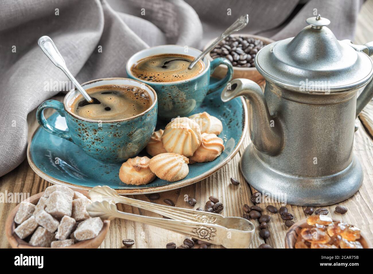Due tazze blu di caffè nero, biscotti e caffettiera circondate da tela di lino, pezzi di zucchero e chicchi di caffè su vecchio tavolo di legno Foto Stock
