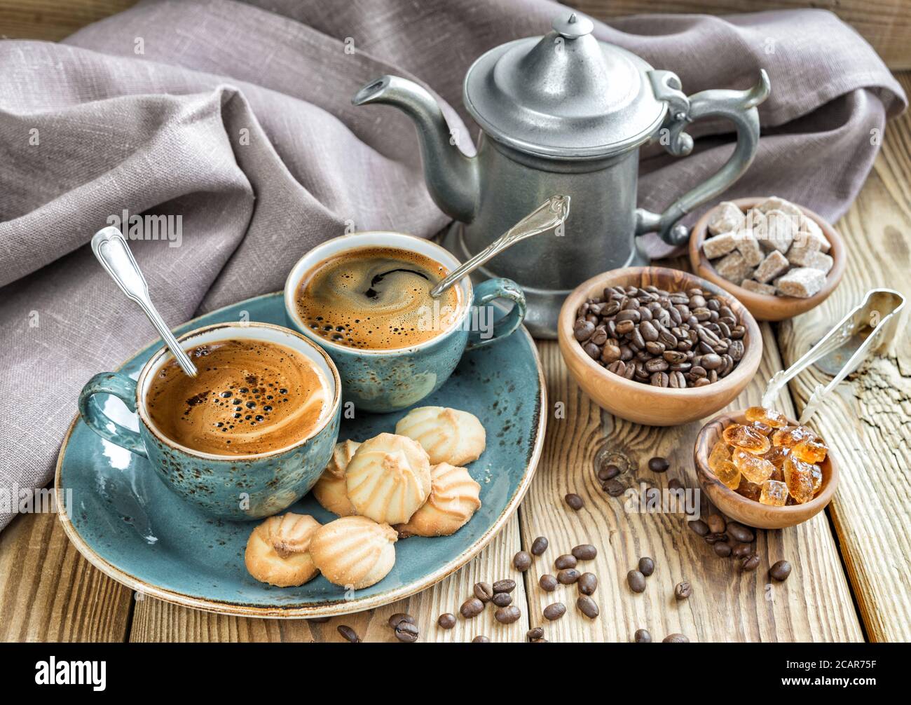 Due tazze blu di caffè nero, biscotti e una caffettiera circondata da tela di lino, zucchero di canna e chicchi di caffè su un vecchio tavolo di legno Foto Stock