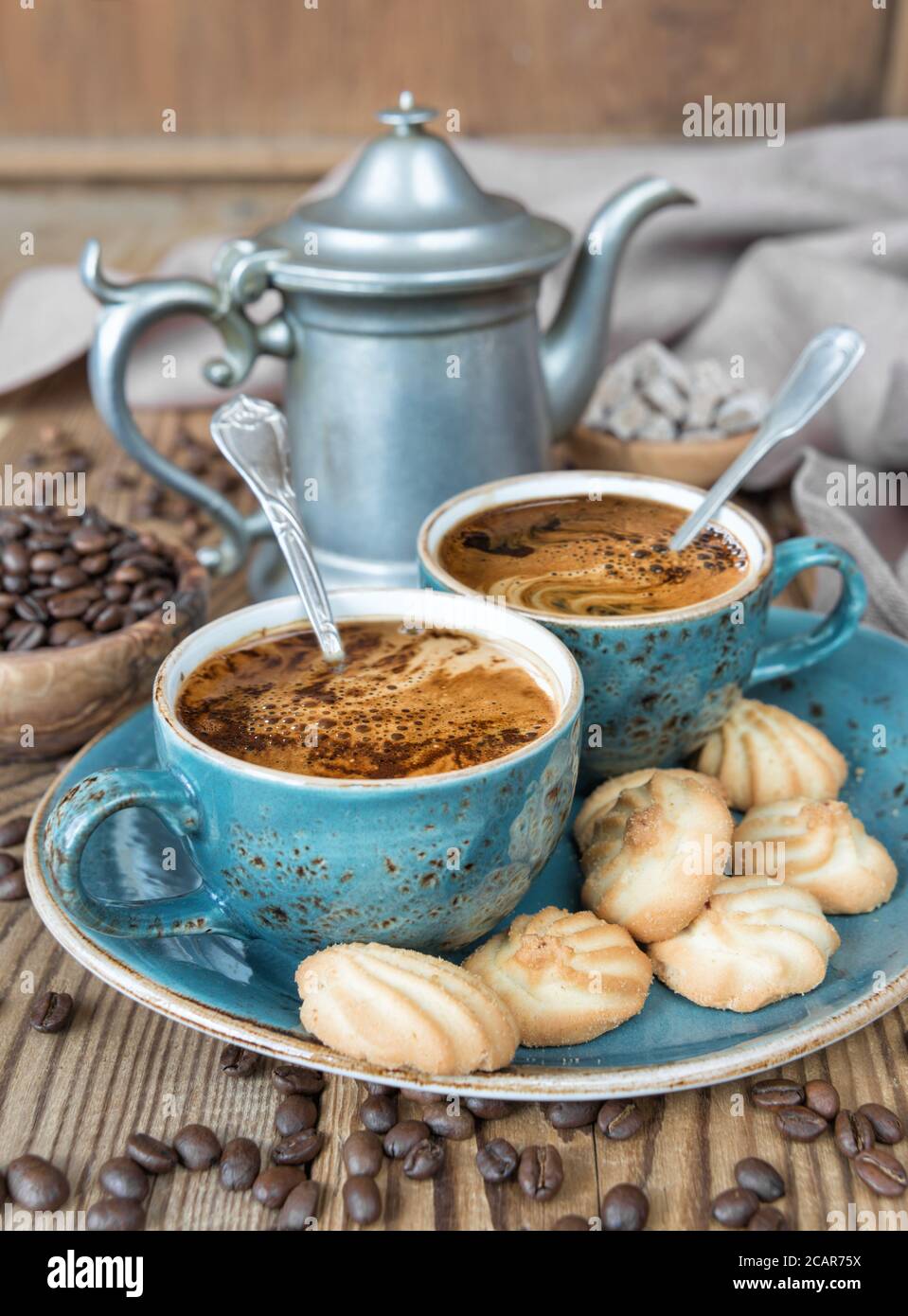 Due tazze blu di caffè nero, biscotti e caffettiera circondate da tela di lino, zucchero di canna e chicchi di caffè sul vecchio tavolo di legno Foto Stock