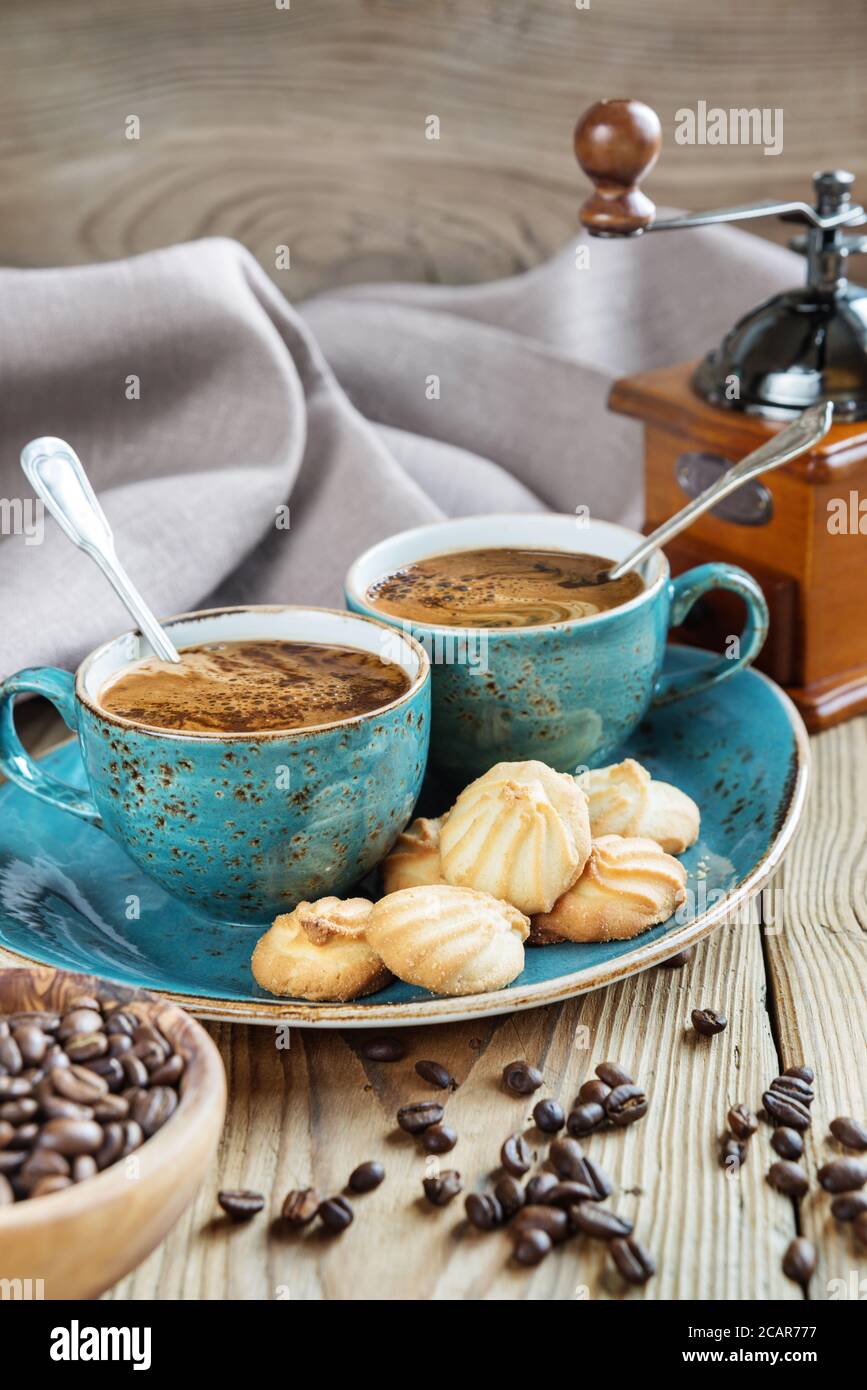 Due tazze blu di caffè nero, biscotti e macinacaffè circondato da tela di lino, pezzi di zucchero e chicchi di caffè sul vecchio tavolo di legno Foto Stock
