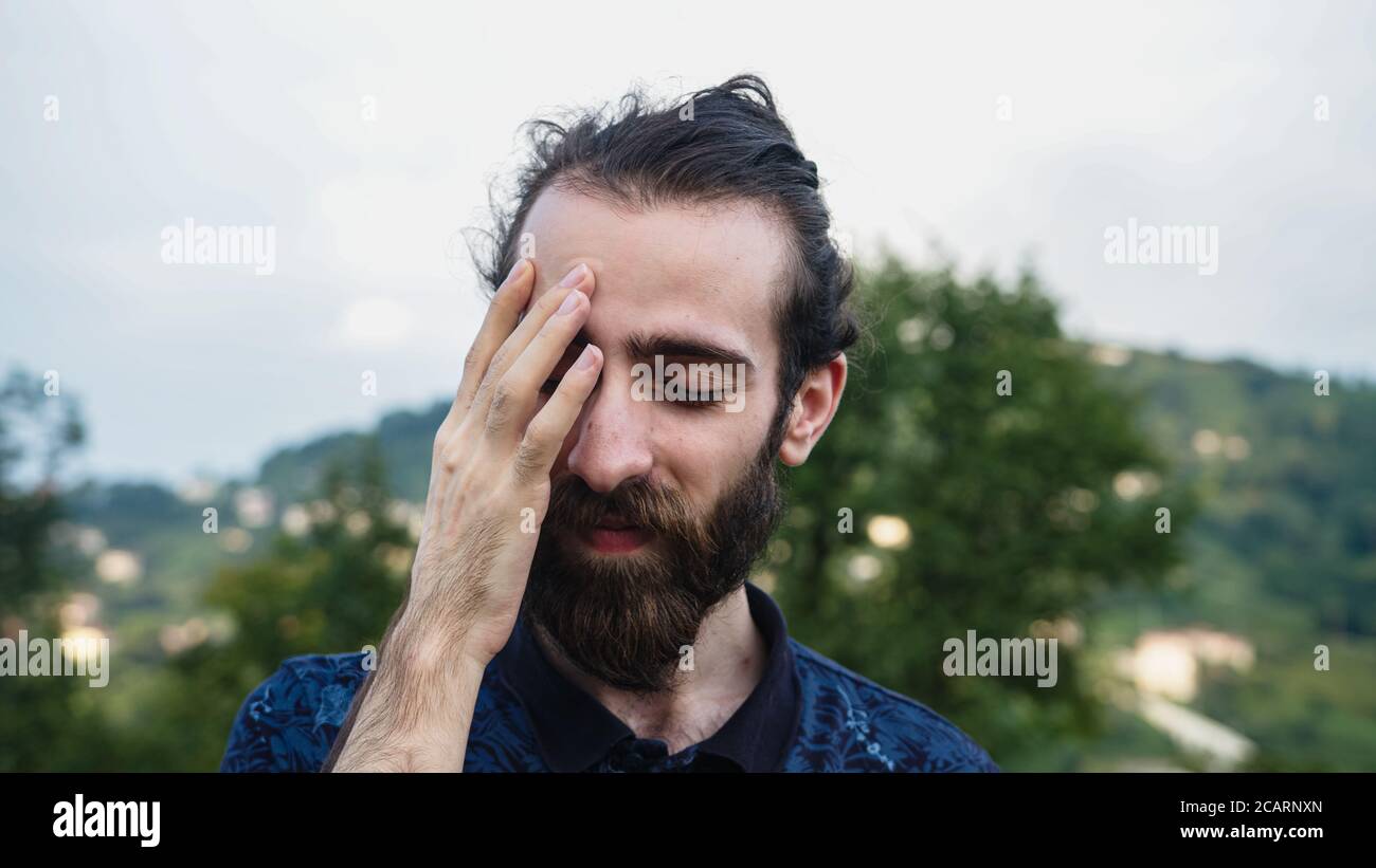 Uomo hipster con capelli lunghi e barba che soffre di mal di testa sembra triste Foto Stock