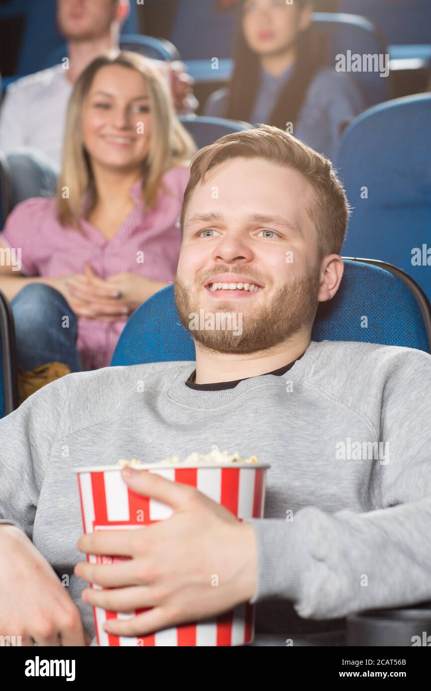 Amante del cinema. Ripresa verticale di un bel giovane bearded che tiene il secchio di popcorn ridendo guardando un film al cinema locale Foto Stock