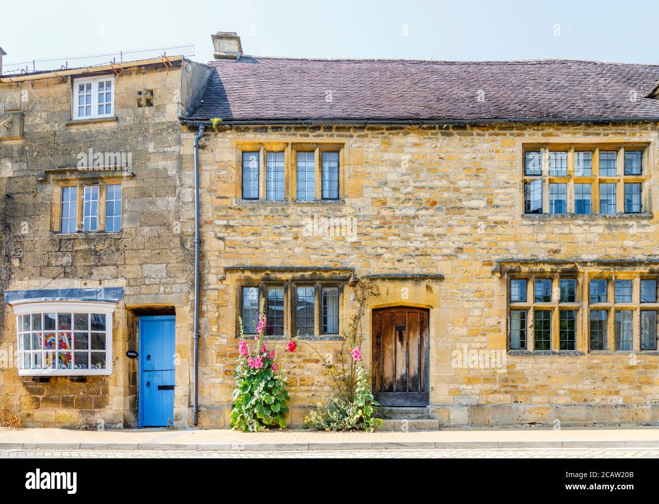 Porta anteriore in legno e bifore in pietra di un cottage con i hollyhocks rosa a rosso che crescono da un muro a Chipping Campden nel Cotswolds Foto Stock