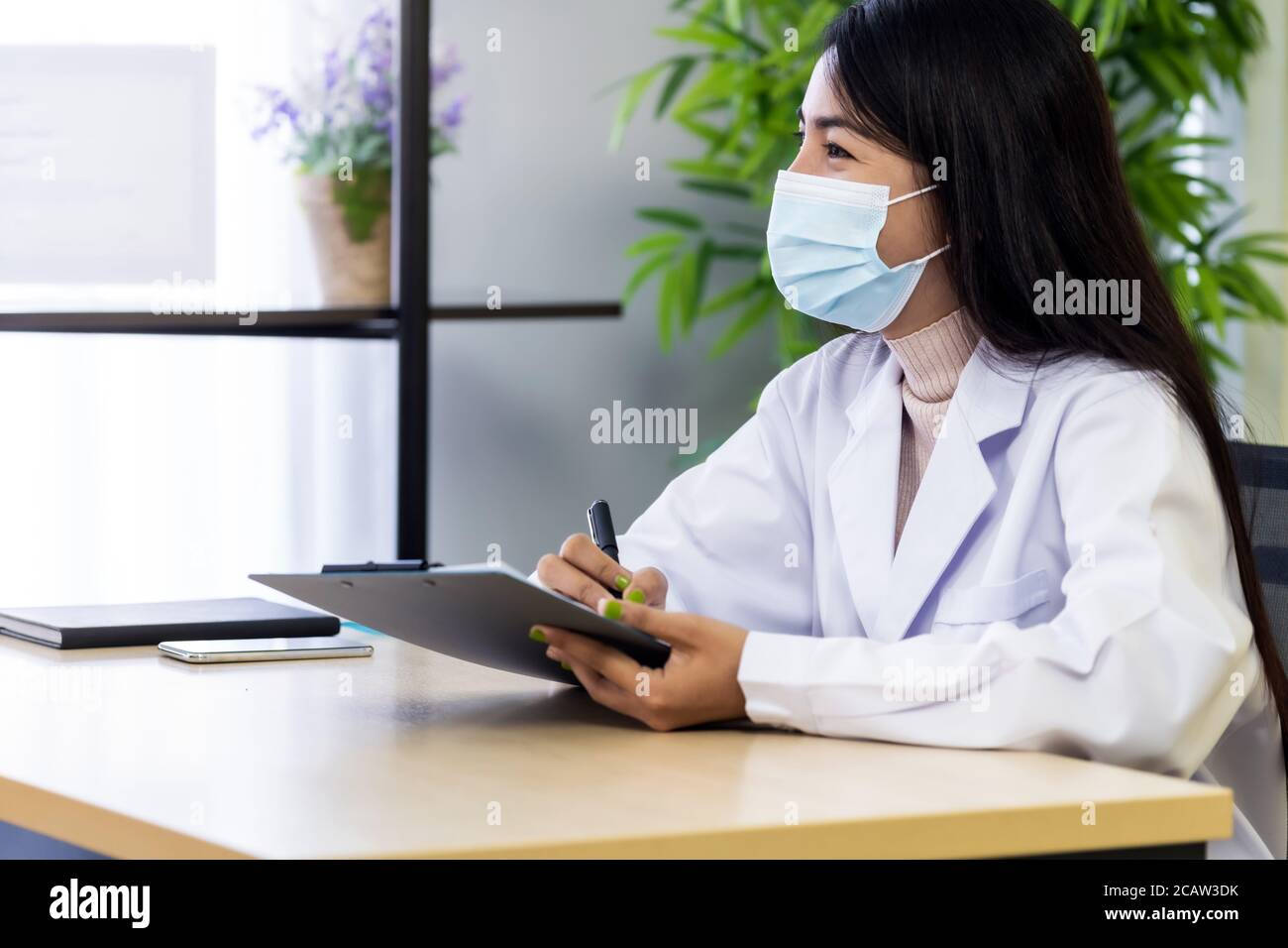 Ritratto di donna asiatica medico indossare maschera protettiva viso sedersi nella sua stanza di ufficio in clinica ospedale e parlare con il paziente durante il controllo del paziente . Est Foto Stock