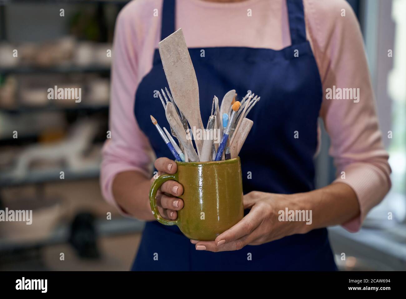 Processo di creazione. Donna artigianale che lavora nella sua officina. Primo piano delle mani femminili che tengono il recipiente in ceramica pieno di spazzole e altri utensili. Negozio di ceramica artigianale, piccola impresa Foto Stock