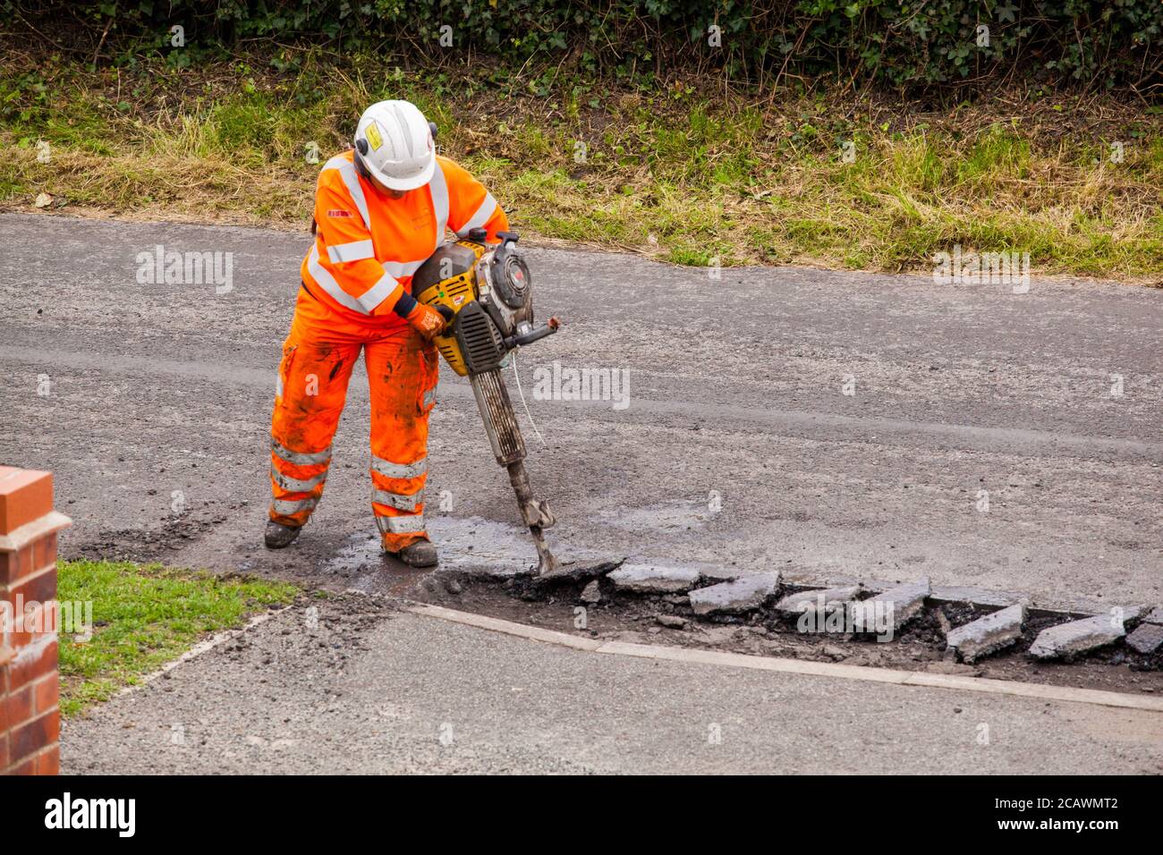 Lavorare la donna con il martello di sollevamento che esegue riparazioni stradali e riparazione dei danni superficie stradale irregolare e fori di pentola con asfalto in a. Cheshire Country Lane Road Foto Stock