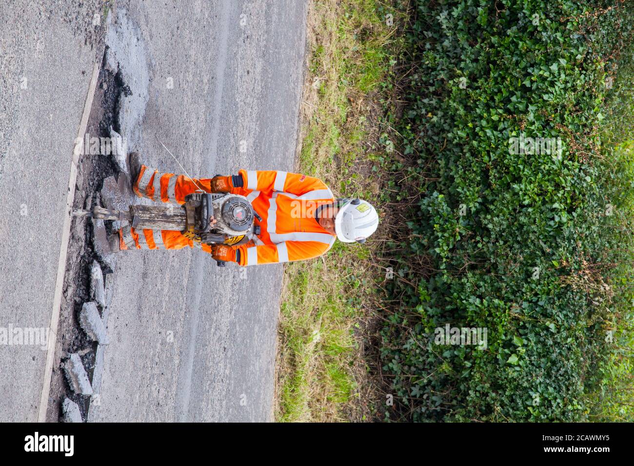 Lavorare la donna con il martello di sollevamento che esegue riparazioni stradali e riparazione dei danni superficie stradale irregolare e fori di pentola con asfalto in a. Cheshire Country Lane Road Foto Stock