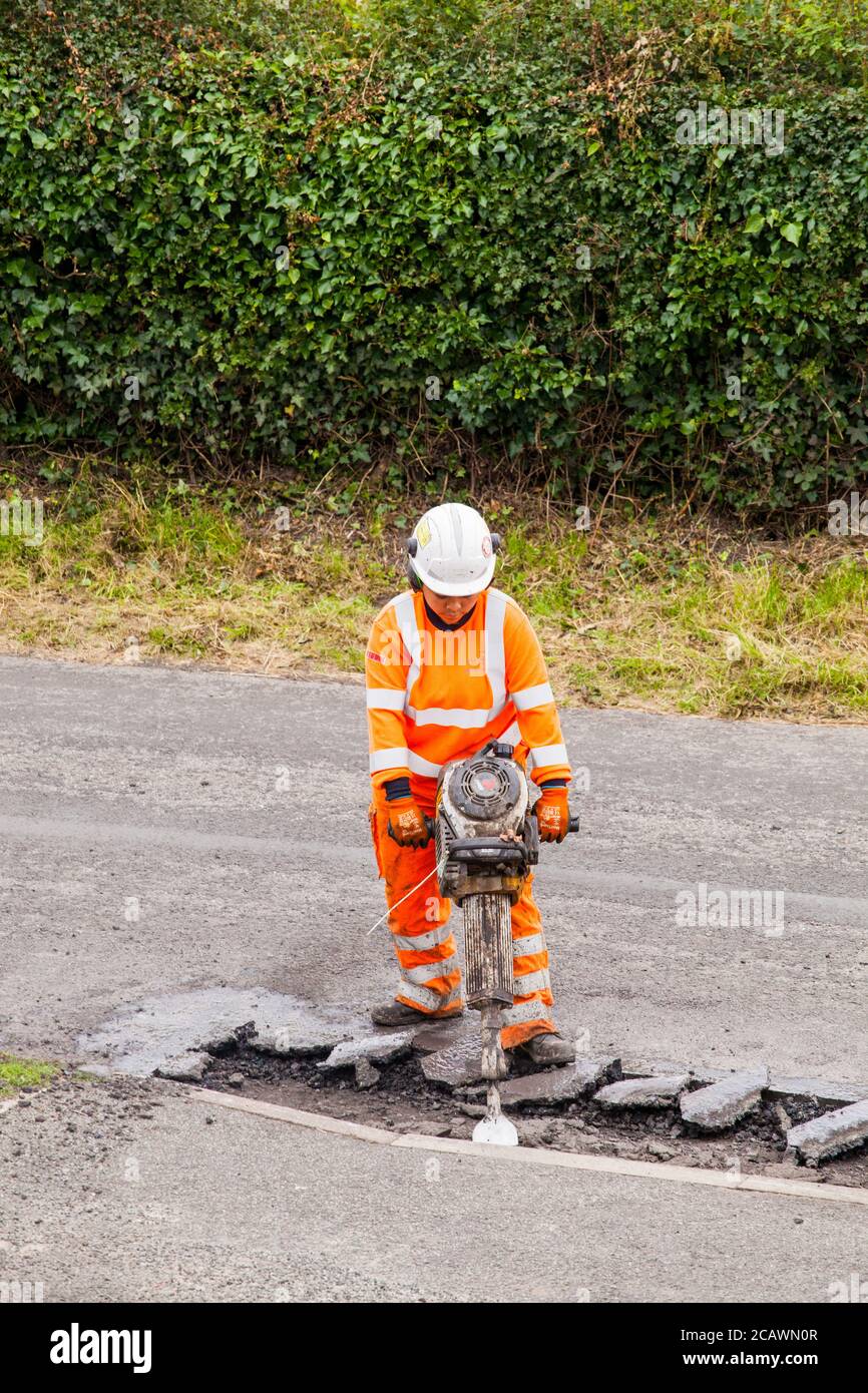 Lavorare la donna con il martello di sollevamento che esegue riparazioni stradali e riparazione dei danni superficie stradale irregolare e fori di pentola con asfalto in a. Cheshire Country Lane Road Foto Stock