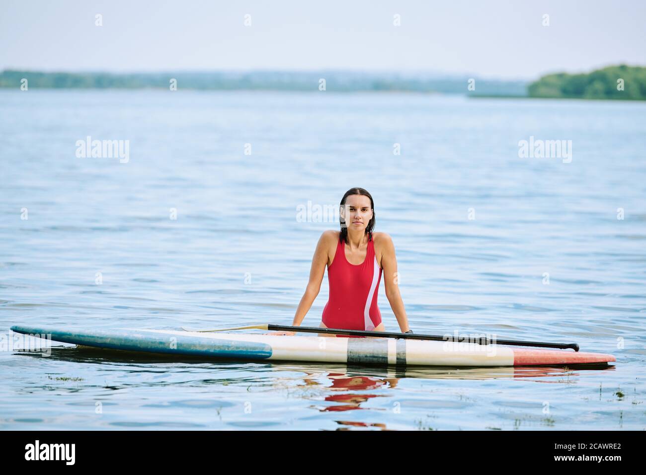 Felice giovane donna bruna in costume da bagno rosso in piedi in acqua con tavola da surf Foto Stock