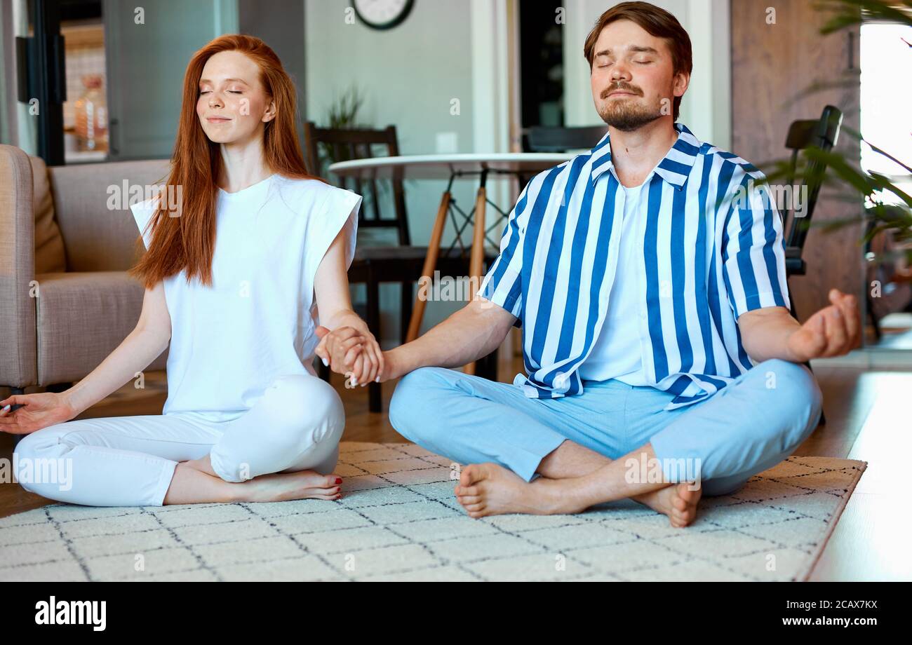 adorabile coppia caucasica meditando a casa, donna rossa e ragazzo bearded in posa yoga, indossando abiti casual. yoga, concetto di stile di vita sano Foto Stock