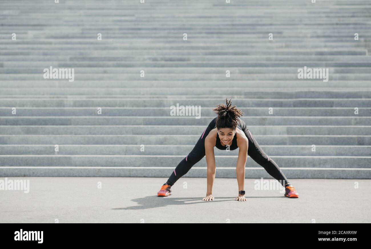 Esercizi per la flessibilità. Gioiosa ragazza afroamericana in abbigliamento sportivo con fitness tracker facendo stretching Foto Stock