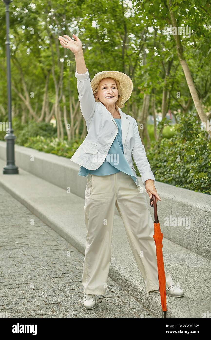 Anziana bianca bella signora di circa 62 anni sta viaggiando mentre in piedi sul marciapiede nel parco pubblico. È cordiale e allegra, vestita in cas Foto Stock