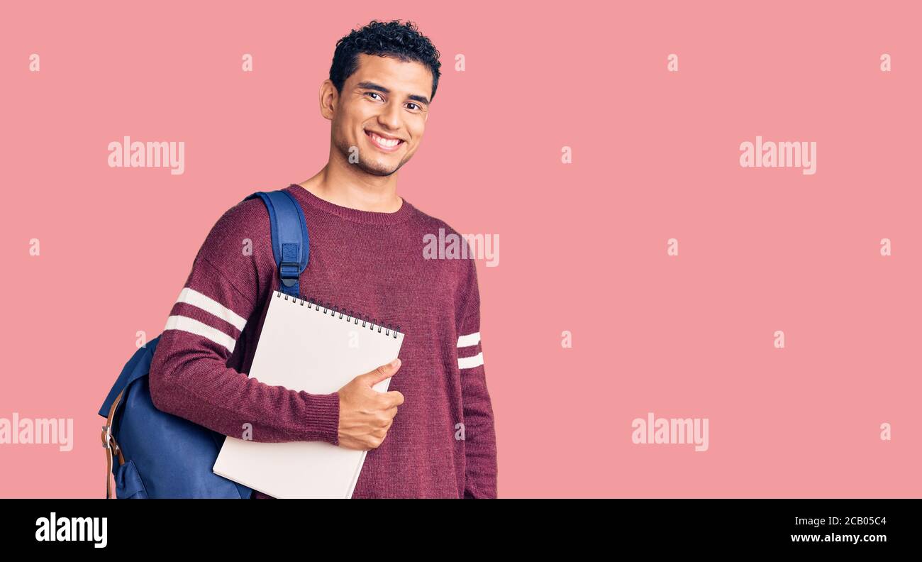 Bell'uomo ispanico che indossa zaino e notebook per studenti che appare positivo e felice in piedi e sorridente con un sorriso sicuro che mostra i denti Foto Stock