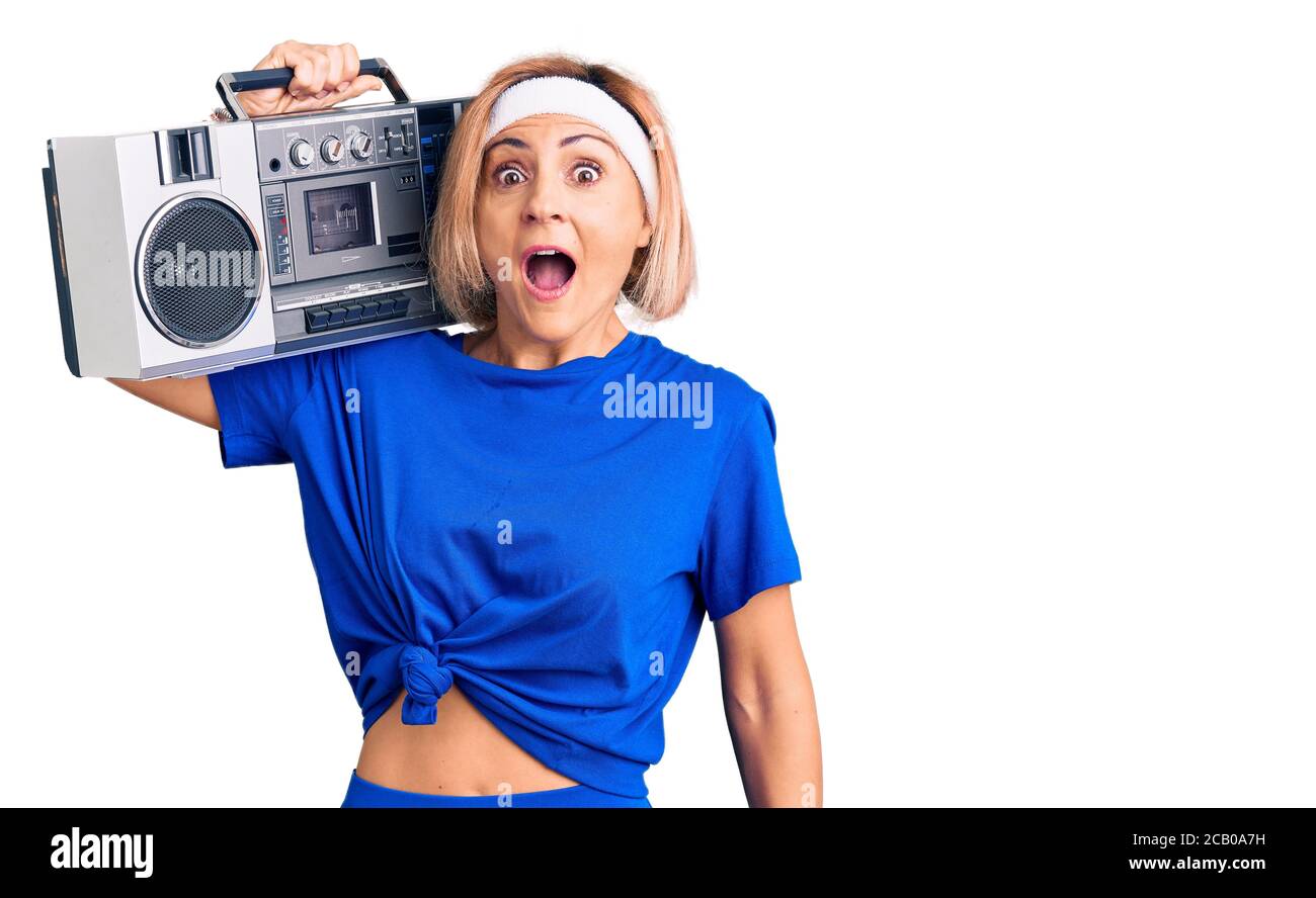 Giovane donna bionda che indossa abbigliamento sportivo reggendo boombox, ascoltando musica spaventato e stupito con bocca aperta per sorpresa, viso incredulo Foto Stock