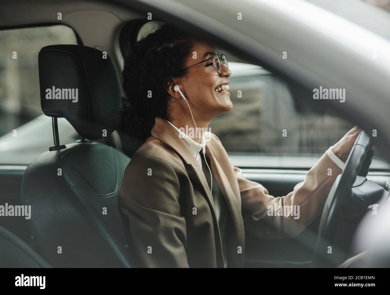 Donna d'affari con gli auricolari che guidano la sua automobile all'ufficio e sorridente. Donna che ama ascoltare la musica mentre guida al lavoro. Foto Stock
