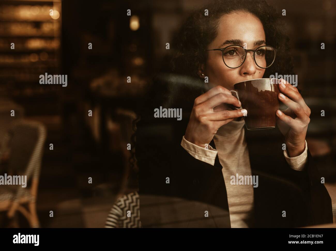 Donna che indossa bicchieri bere caffè e guardare via. Donna seduta all'interno del caffè con una tazza di caffè. Foto Stock