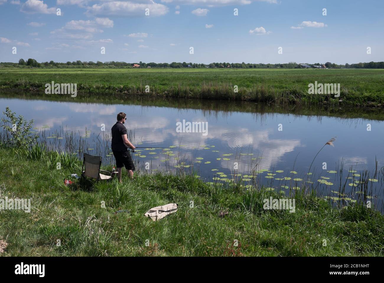 Il pescatore con la sigaretta in bocca si alza con un bastone da pesca sulla riva di un piccolo fiume. Nuvole si specchiavano in acqua nel tipico paesaggio olandese Foto Stock