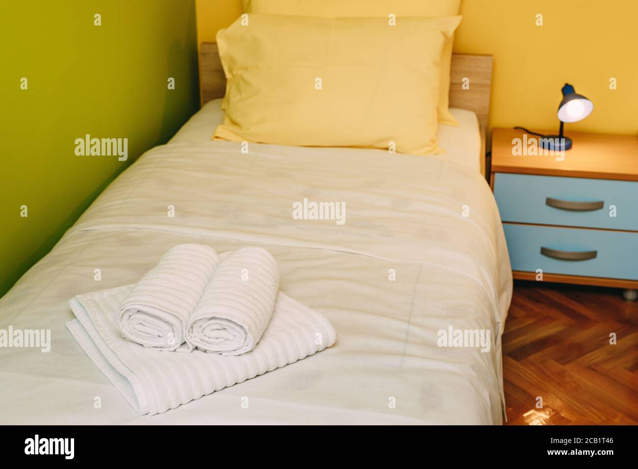 Un letto singolo con cuscini gialli e una coperta bianca in una camera con pareti gialle e verdi, un comodino di legno blu con una lampada da lettura e terry Foto Stock