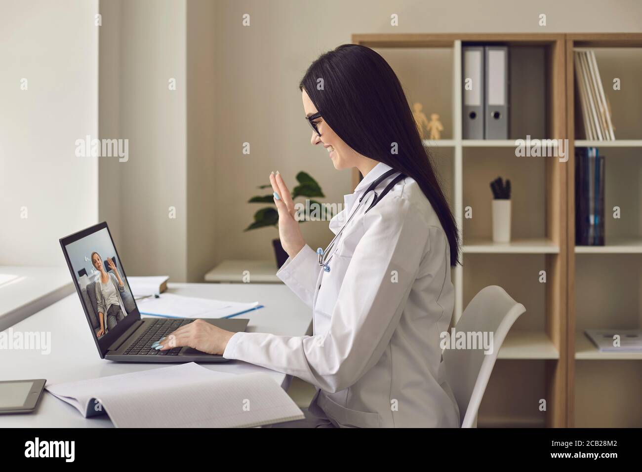 Donna dottore nel suo ufficio con un computer portatile che si sventola al paziente. Il medico in linea dà la consultazione remota usando la videochiamata alla donna anziana del paziente. Foto Stock